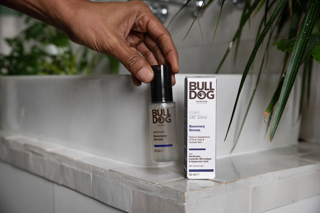 A hand holds an End of Day Recovery Serum bottle, formulated to target fine lines, beside its packaging on a white bathroom sink with green plants nearby.