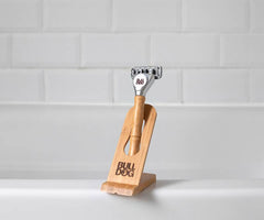 A Bulldog Bamboo Razor with a natural bamboo handle stands upright on the Bamboo Razor Stand, both featuring the BULL DOG logo, set against a white tiled background.