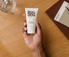 A hand holds a tube of Original Hand Cream on a wooden table beside a glass of water and a brown notebook with a white booklet; made with natural origin ingredients for moisturizing care.