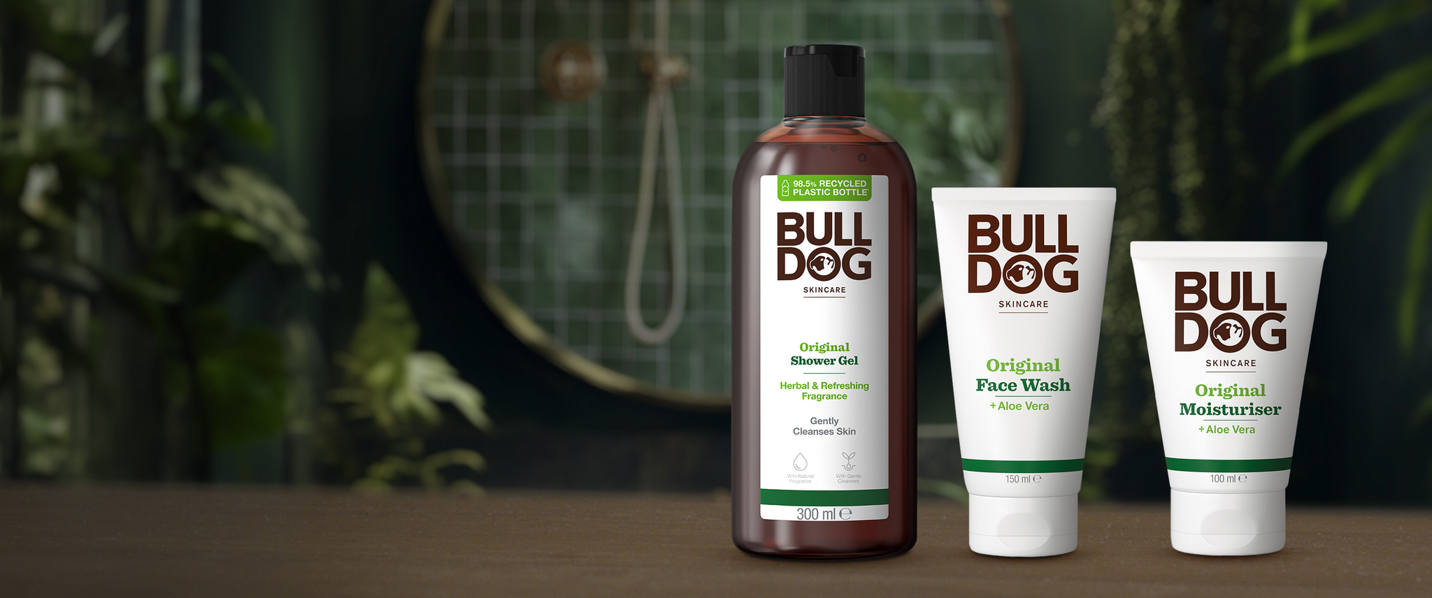 Three Bulldog Skincare products—Original shower gel, face wash, and moisturiser—are displayed in front of a green tiled background with a circular mirror partially visible.