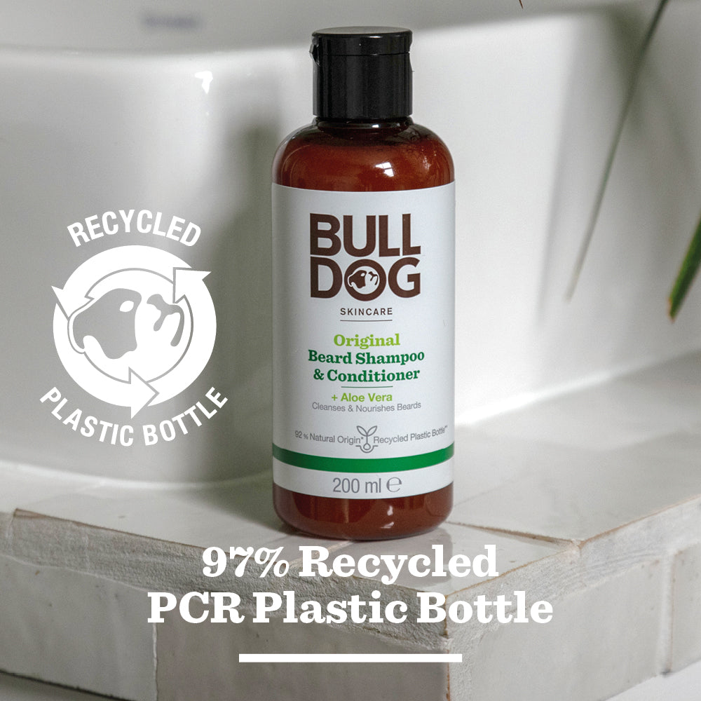 A bottle of Original Beard Shampoo and Conditioner rests on a bathroom counter, its label featuring natural origin ingredients and eco-friendly packaging made from 97% recycled PCR plastic, plus a visible recycling symbol.