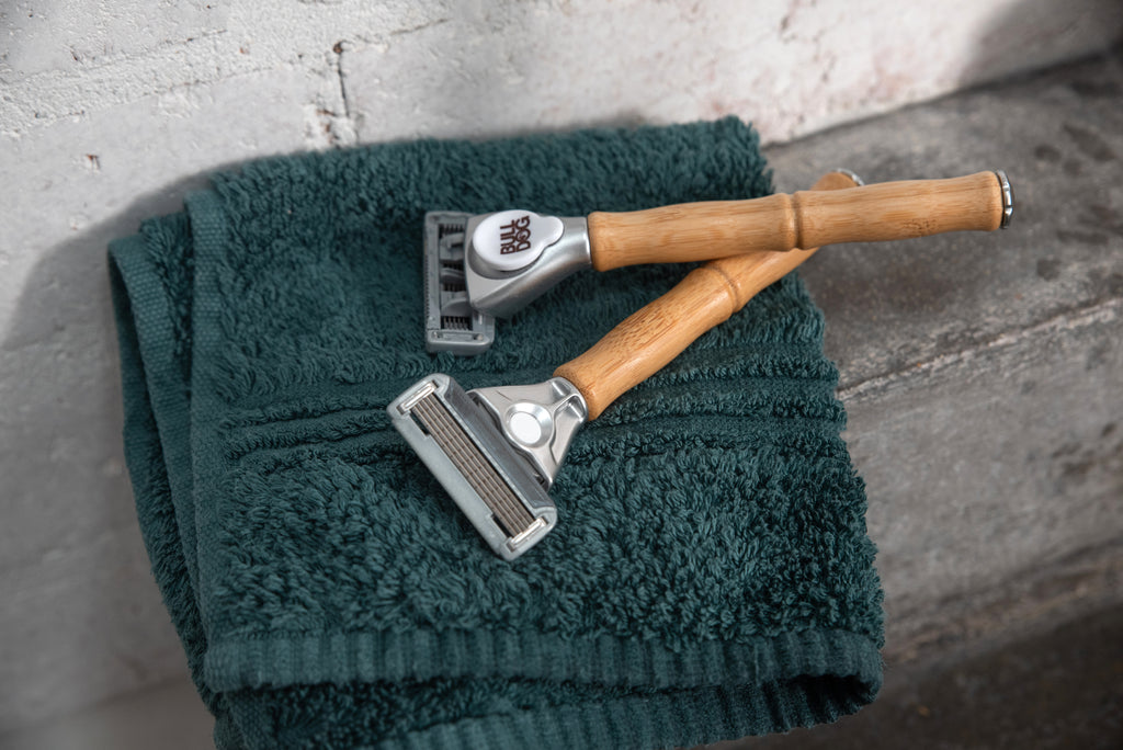 Two Original Steel Blades razors with bamboo handles rest on a folded dark green towel atop a stone or concrete surface beside a white textured wall.
