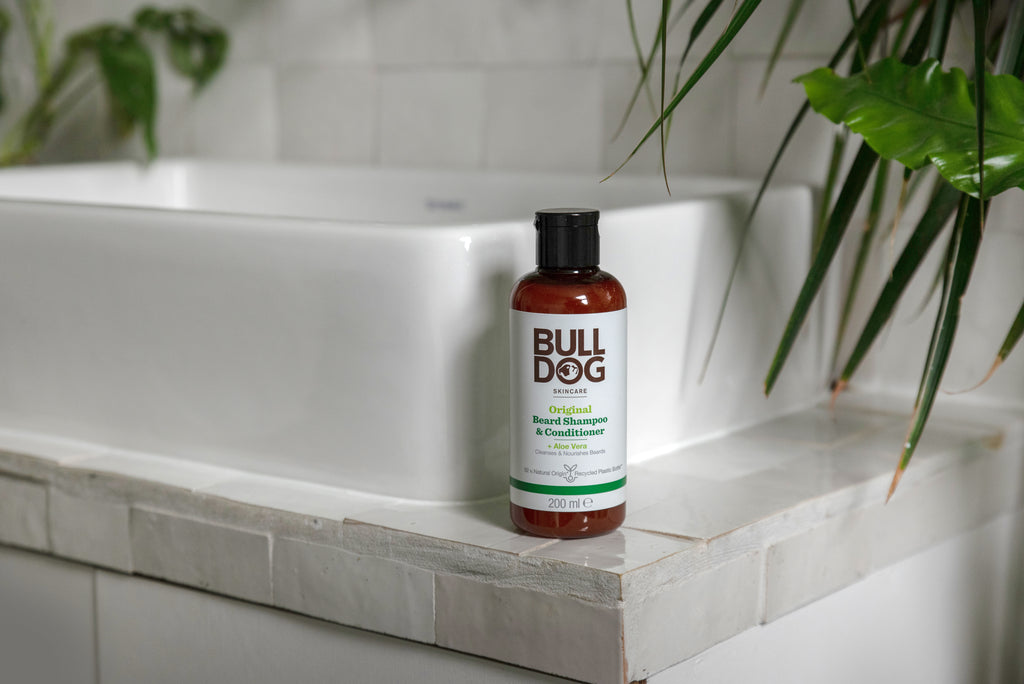 A bottle of Original Beard Shampoo and Conditioner, made with natural origin ingredients, sits on the edge of a white tiled bathroom sink with green plants nearby.