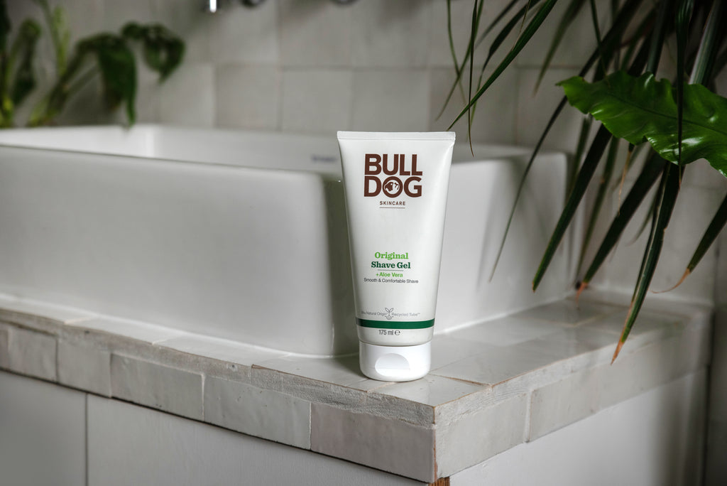 A tube of Original Shave Gel sits on the edge of a white bathroom sink with green plants nearby, emphasizing its natural origin ingredients such as soothing aloe vera.
