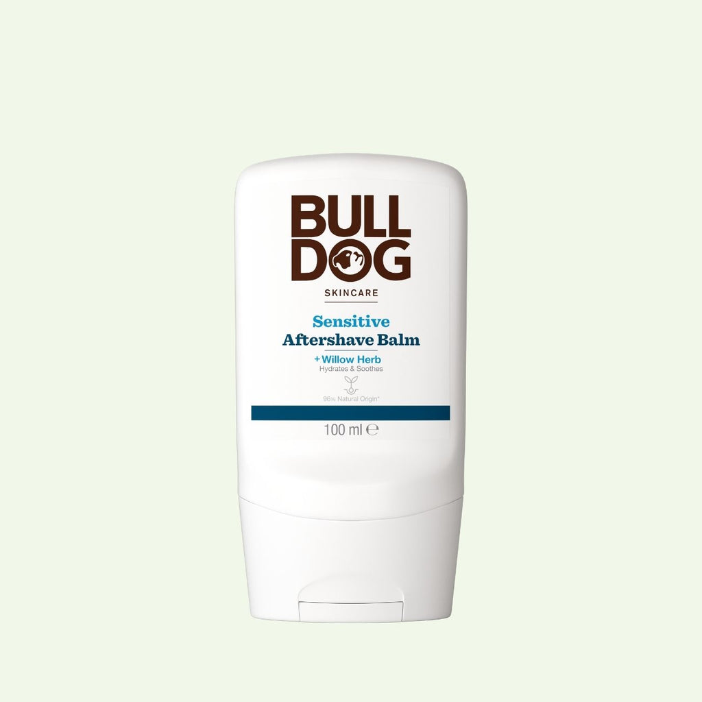 A white 100ml bottle of Bulldog Skincare Sensitive Aftershave Balm, designed for sensitive skin and made with natural origin ingredients, featuring bold brown and blue text, is set against a pale green background.