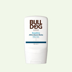 A white 100ml bottle of Bulldog Skincare Sensitive Aftershave Balm, designed for sensitive skin and made with natural origin ingredients, featuring bold brown and blue text, is set against a pale green background.