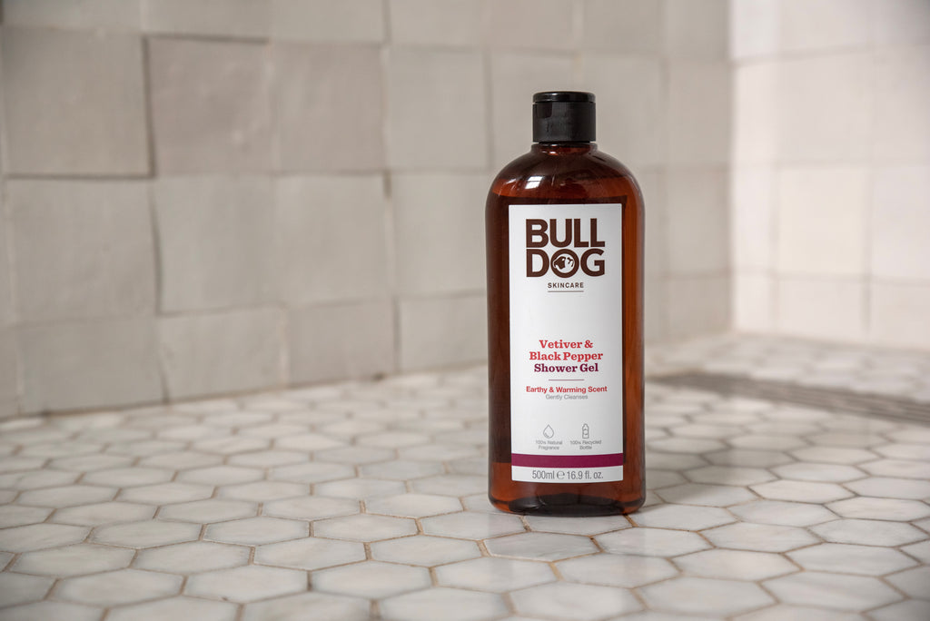 A bottle of Vetiver & Black Pepper Shower Gel, designed for men, stands on a white hexagon-tiled surface. The brown bottle has a black cap and white label, placed against a tiled wall backdrop.
