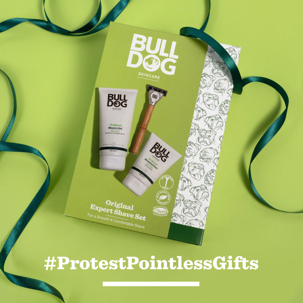 An Expert Shave Trio by Bulldog Skincare for Men, featuring a bamboo razor, is displayed on a green surface with a ribbon and #ProtestPointlessGifts in bold white text below.