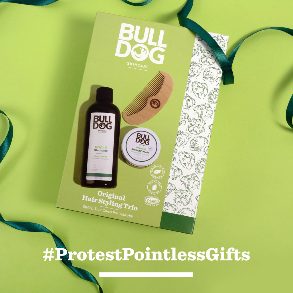 A Bulldog Skincare Hair Styling Trio gift set, including men's hair care products and a wooden comb, rests on a green background with a ribbon. Text below reads #ProtestPointlessGifts—ideal for men who value quality.