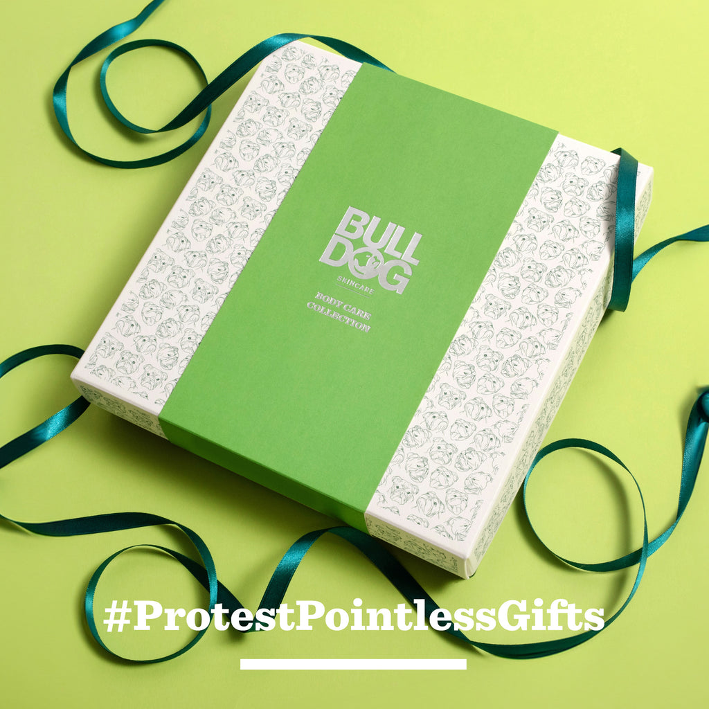 A green and white Premium Body Care Collection gift box rests on a light green surface with scattered blue ribbon. Text reads: #ProtestPointlessGifts—perfect for those seeking thoughtful gifts for men.
