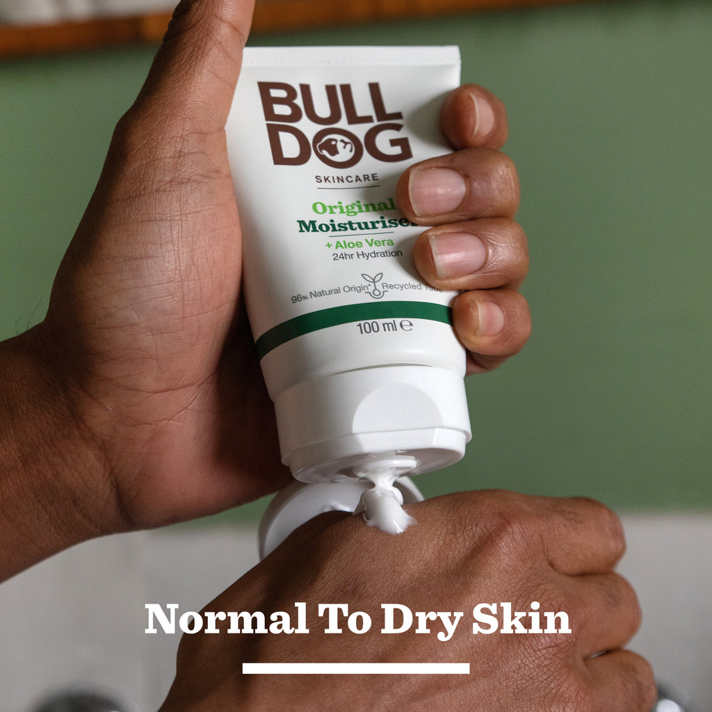 A person squeezes Bulldog Skincare Original Grooming Kit moisturiser onto their hand. The text reads "Normal To Dry Skin," highlighting this kit as a perfect grooming essential for men. The background appears slightly blurred.
