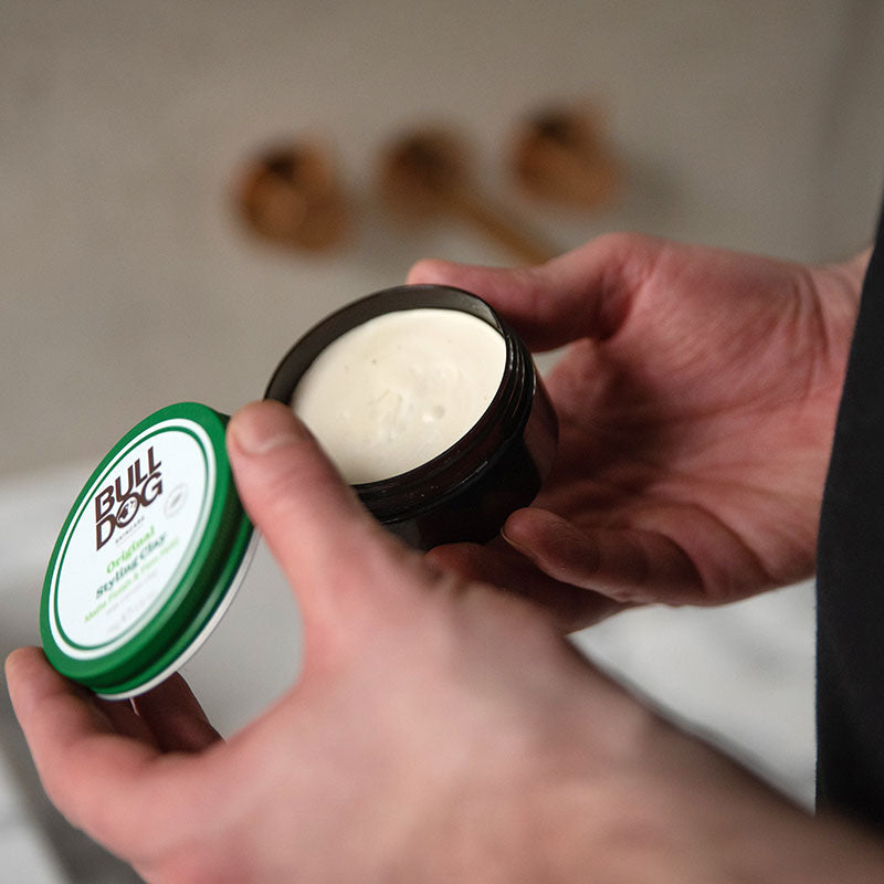 A person holds an open jar of Bulldog Original Hair Styling Clay, displaying the creamy product inside—perfect for achieving a smooth, matte finish and balanced style. The green and white lid is held in their other hand.
