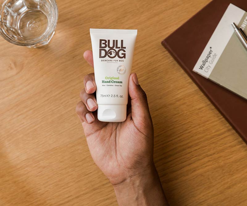 A hand holds a tube of Original Hand Cream on a wooden table beside a glass of water and a brown notebook with a white booklet; made with natural origin ingredients for moisturizing care.